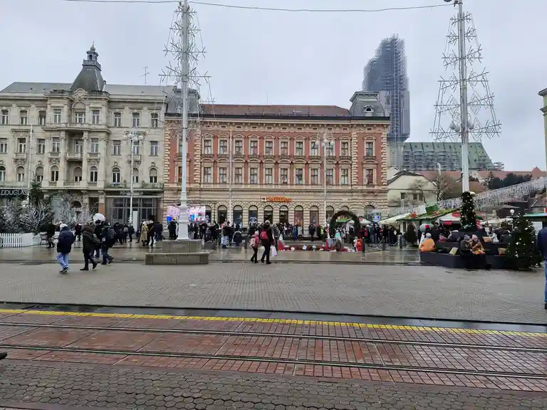 A wide shot of a busy European city square decorated for Christmas with large metal tree frames, festive lights, and crowds…