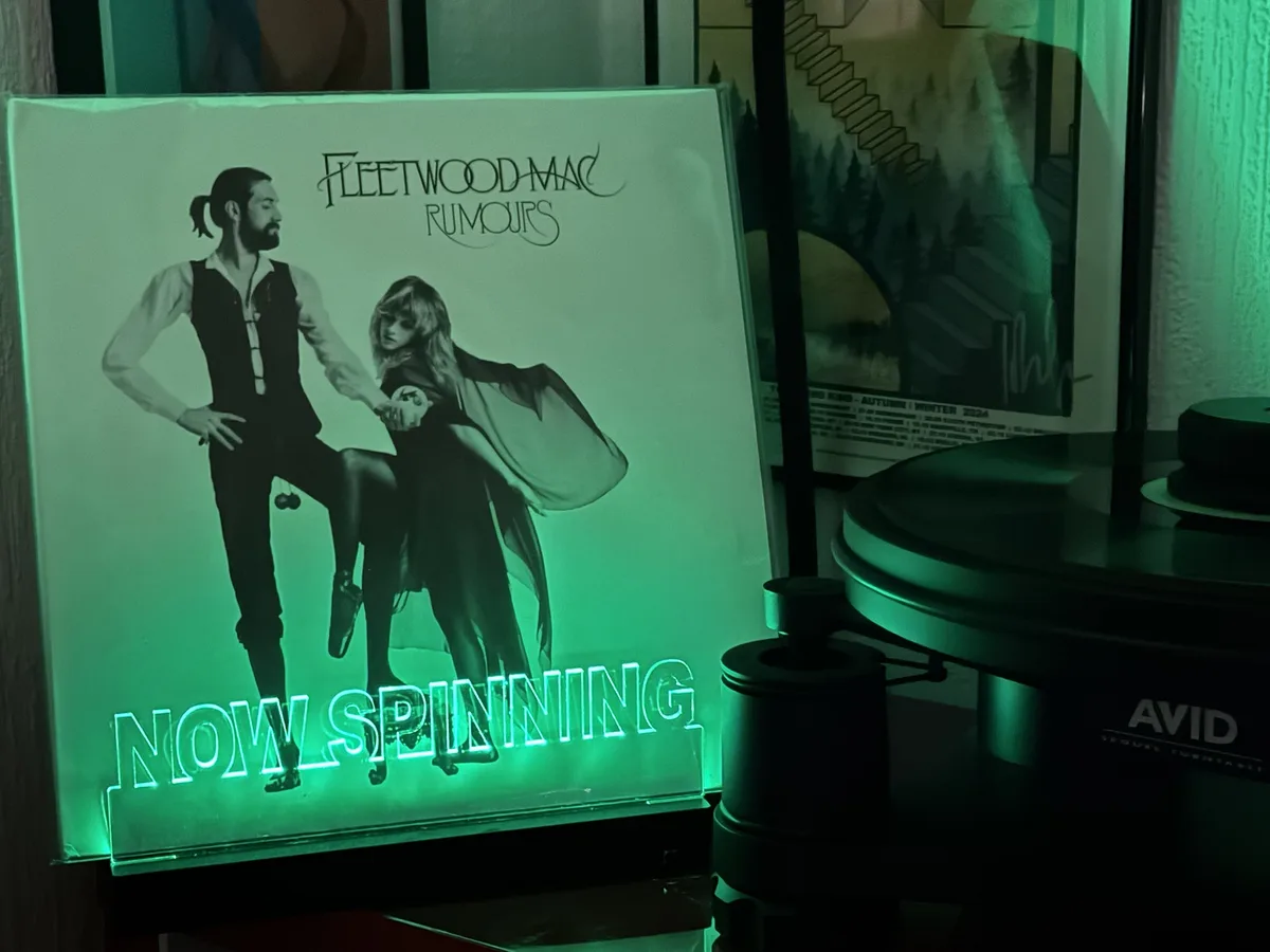 A green-lit, illuminated display of the Fleetwood Mac 'Rumours' album cover stands next to an AVID turntable. The glowing…