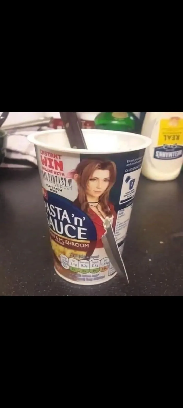 A close-up photo of a cup of instant pasta and sauce with a character from Final Fantasy VII on the label, with a knife…