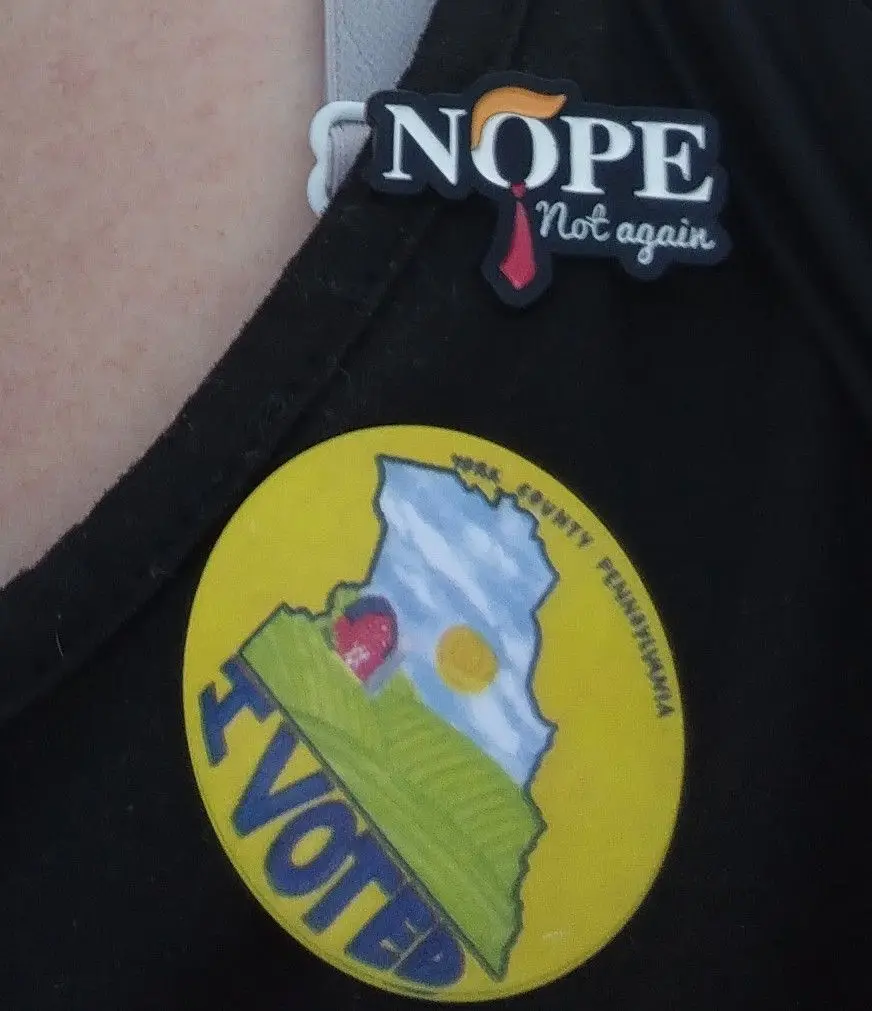 A close-up of a black shirt with two pins: a circular yellow 'I VOTED' pin featuring a map of York County, Pennsylvania, and…