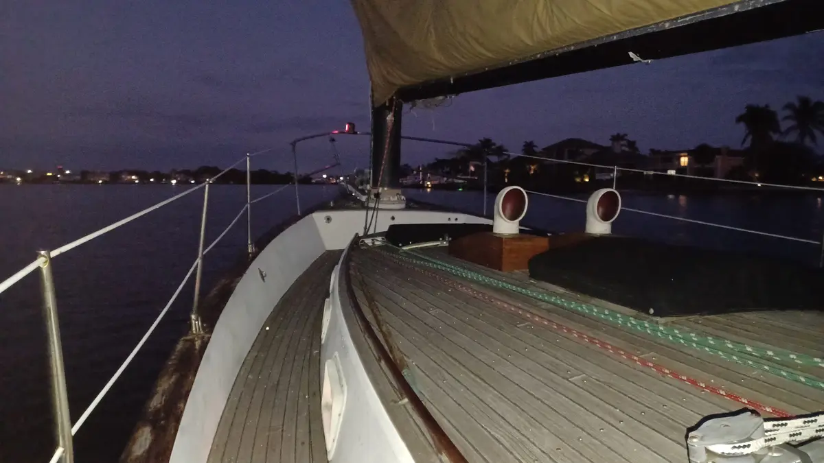A nighttime photo from the deck of a sailboat, showing wooden planks, railings, and distant shoreline lights under a dark…