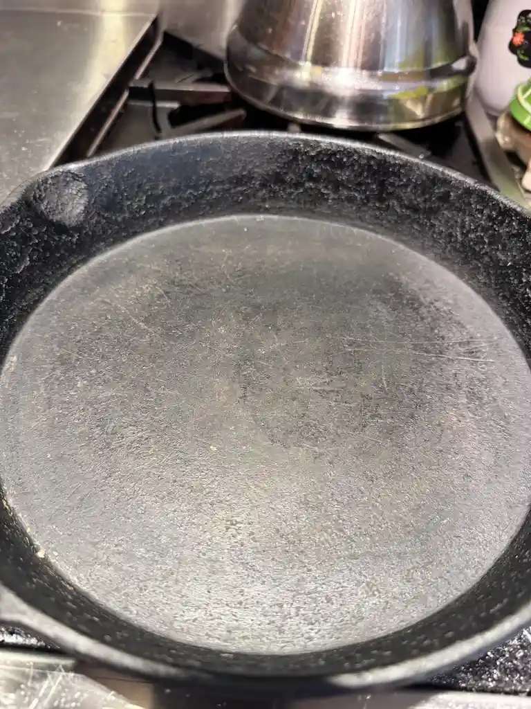 A close-up photo shows a well-used, black cast iron skillet sitting on a stovetop, with visible scratches and wear on its…