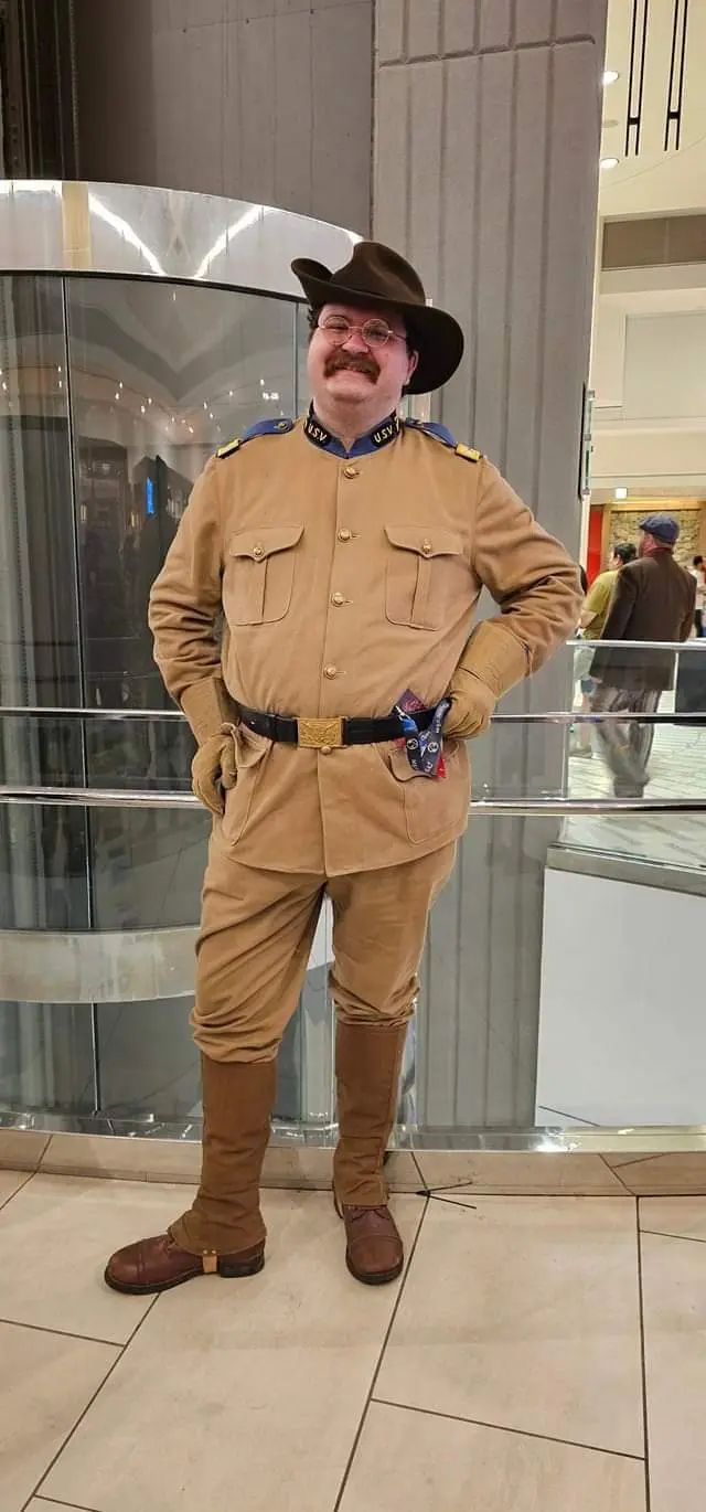A man in a tan military-style uniform with a brown hat and glasses stands confidently in a modern mall, hands on hips. The…