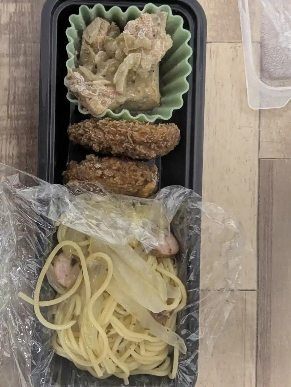 A black rectangular takeout container holds a meal with spaghetti topped with plastic wrap, two fried items, and a green…