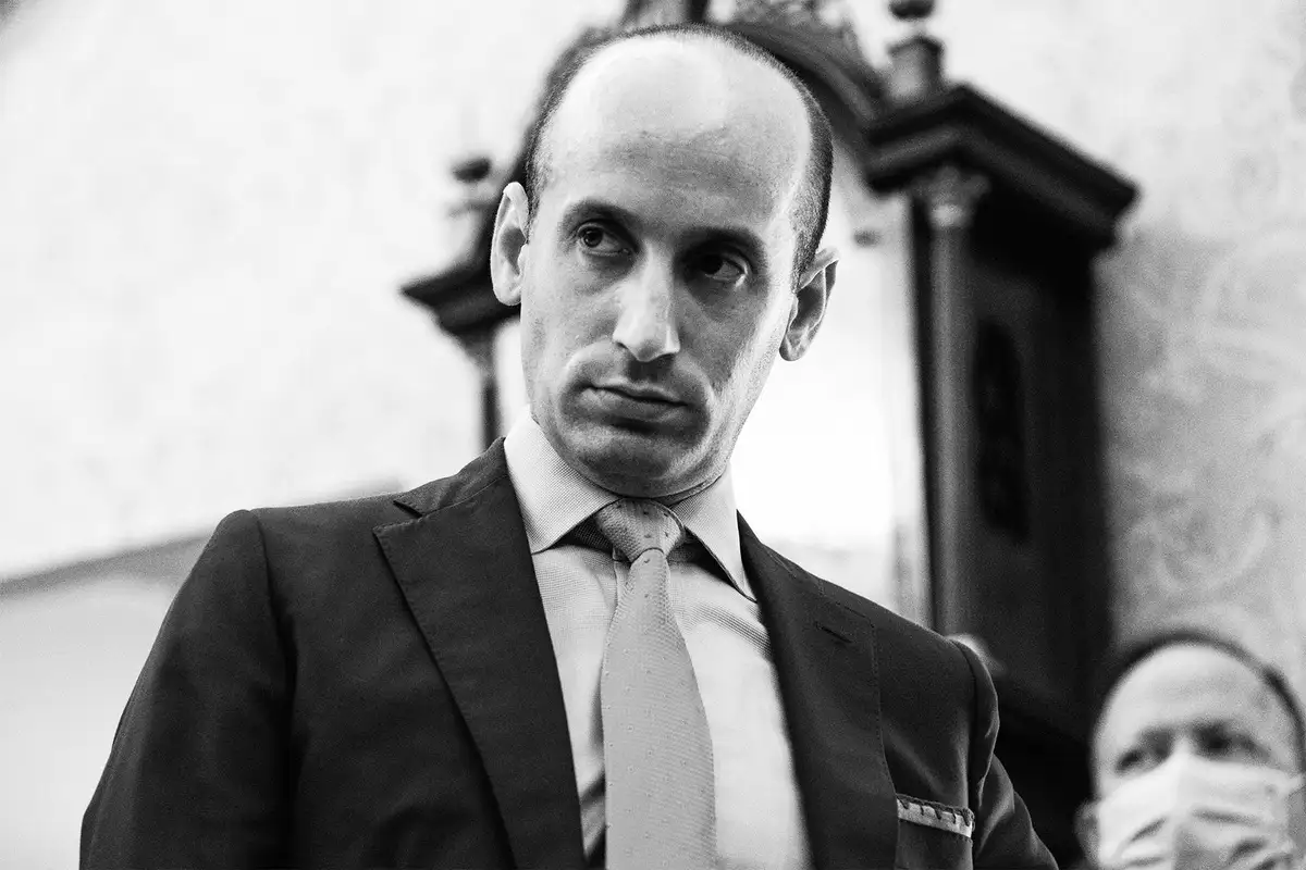 A black-and-white photo shows a man in a suit and tie looking off to the side with a confused or skeptical expression…