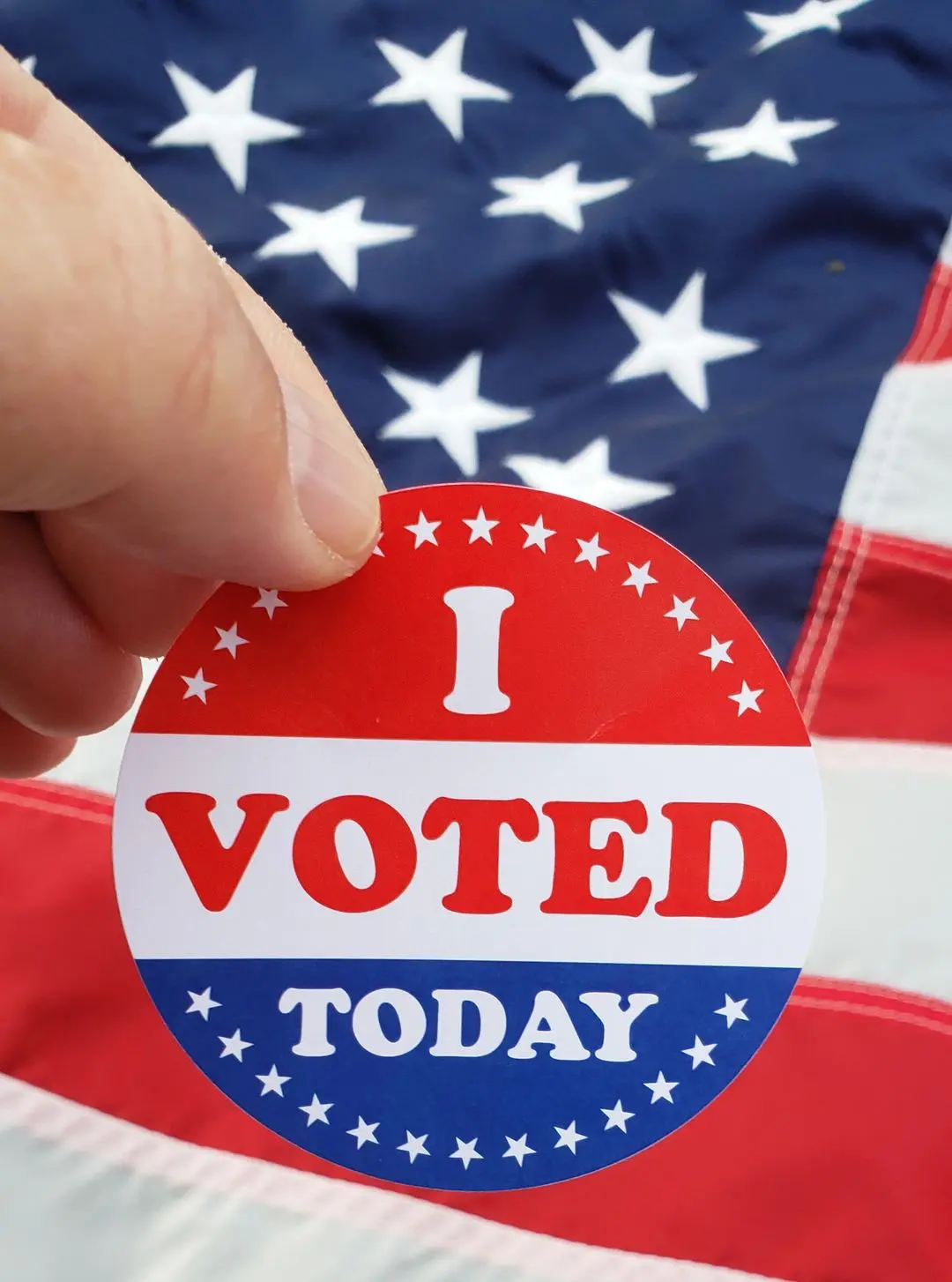 A close-up photo shows a person's thumb holding a circular red, white, and blue 'I VOTED TODAY' sticker against a blurred…