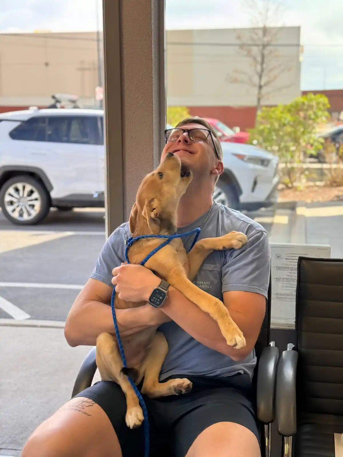 A man wearing glasses and a grey t-shirt sits in a black office chair indoors, holding a light brown puppy in his arms. The…