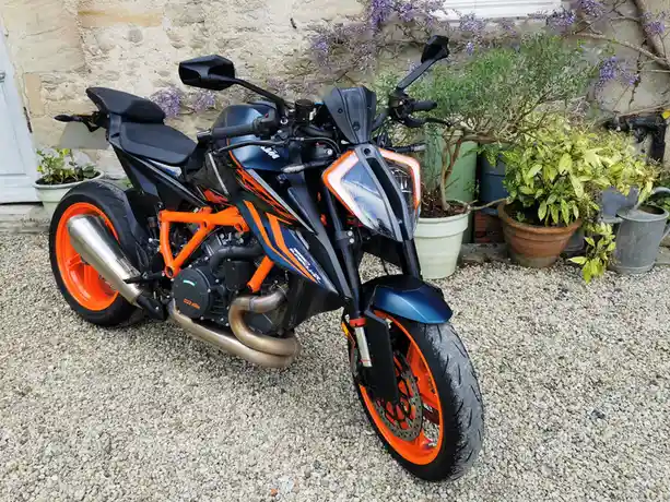 A black and orange KTM motorcycle with visible engine and exhaust pipes is parked on gravel in front of a stone wall with…