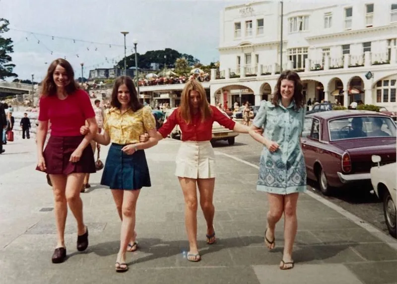four young women from the 1970s walk arm-in-arm down a city street, smiling at the camera. They wear colorful, patterned…