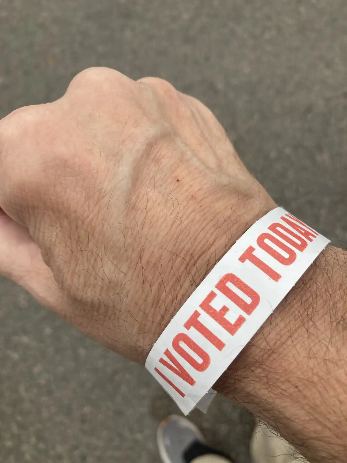 A close-up photo shows a person's hairy, wrinkled hand with a white paper wristband that reads 'I VOTED TODAY!' in bold red…
