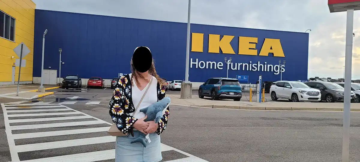 A person stands in a crosswalk in front of an IKEA store, holding a blue plush shark. The store's iconic blue and yellow…