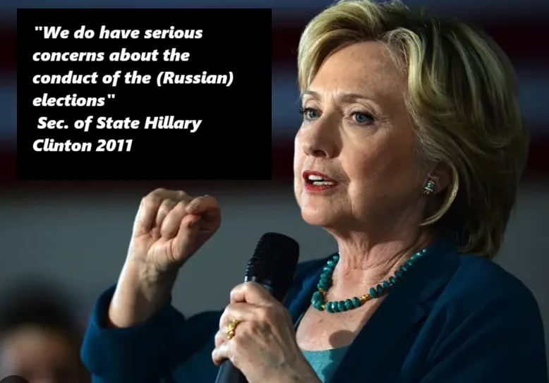 A photo of Hillary Clinton speaking into a microphone, wearing a blue blazer and turquoise necklace, with her right hand…