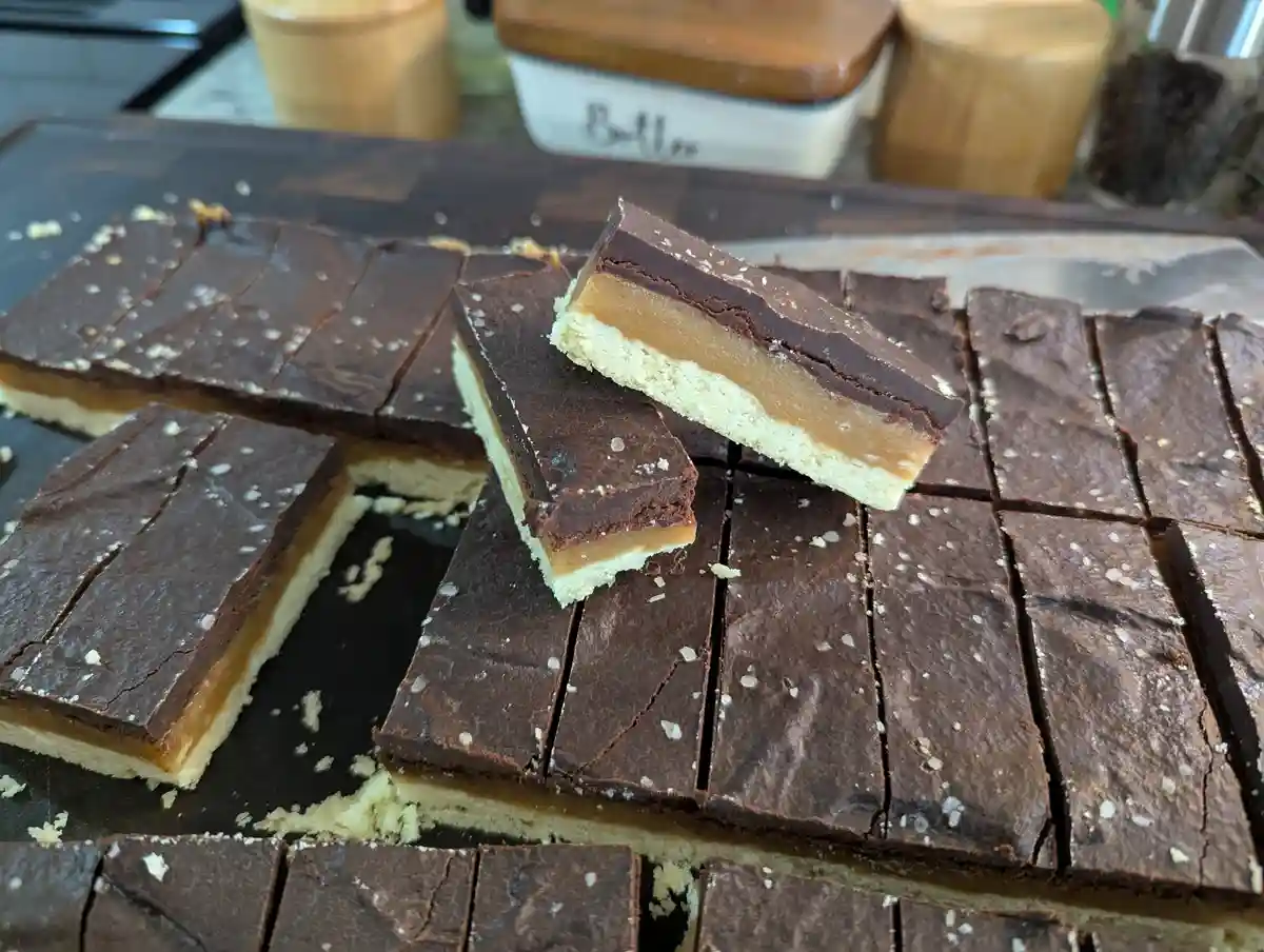 A close-up photo shows homemade chocolate caramel bars cut into squares on a dark cutting board, with one piece lifted to…