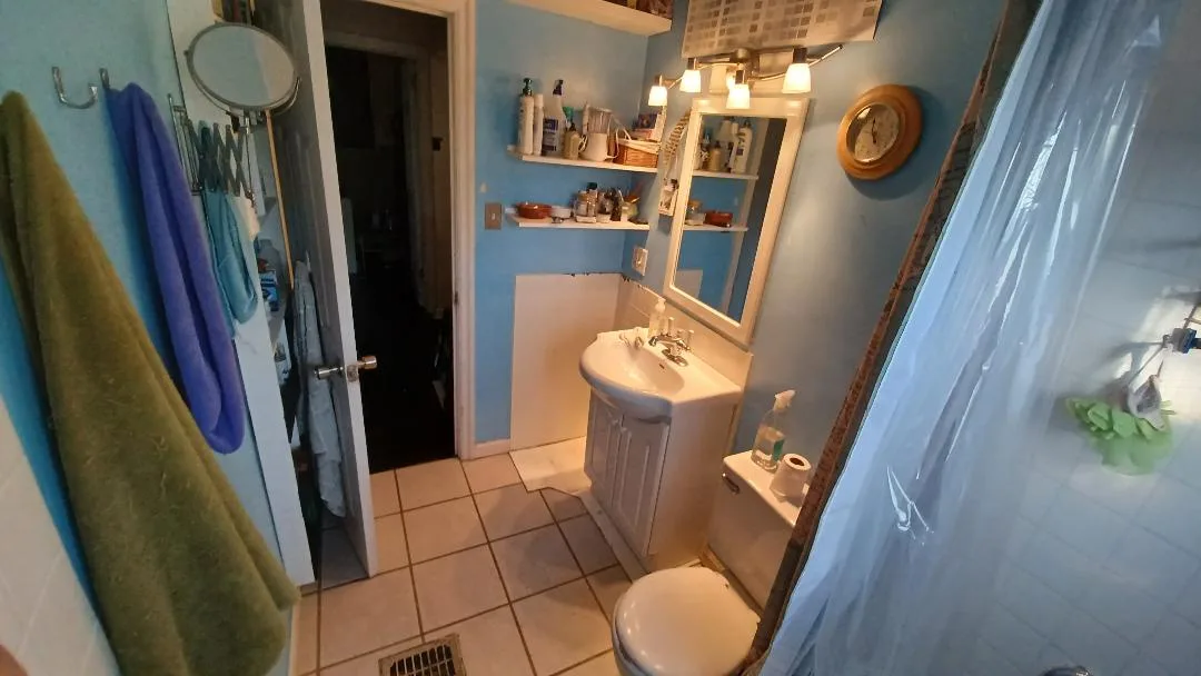 A photograph of a small, cluttered bathroom with light blue walls, a white sink cabinet, toilet, and shower curtain. Towels…