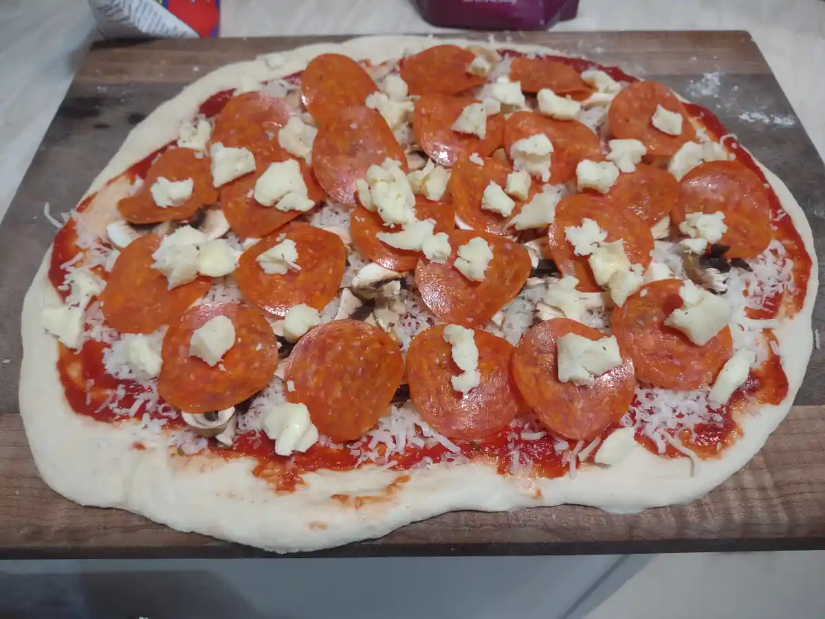 An uncooked pizza on a wooden board has a base of dough, tomato sauce, shredded cheese, sliced mushrooms, and numerous…