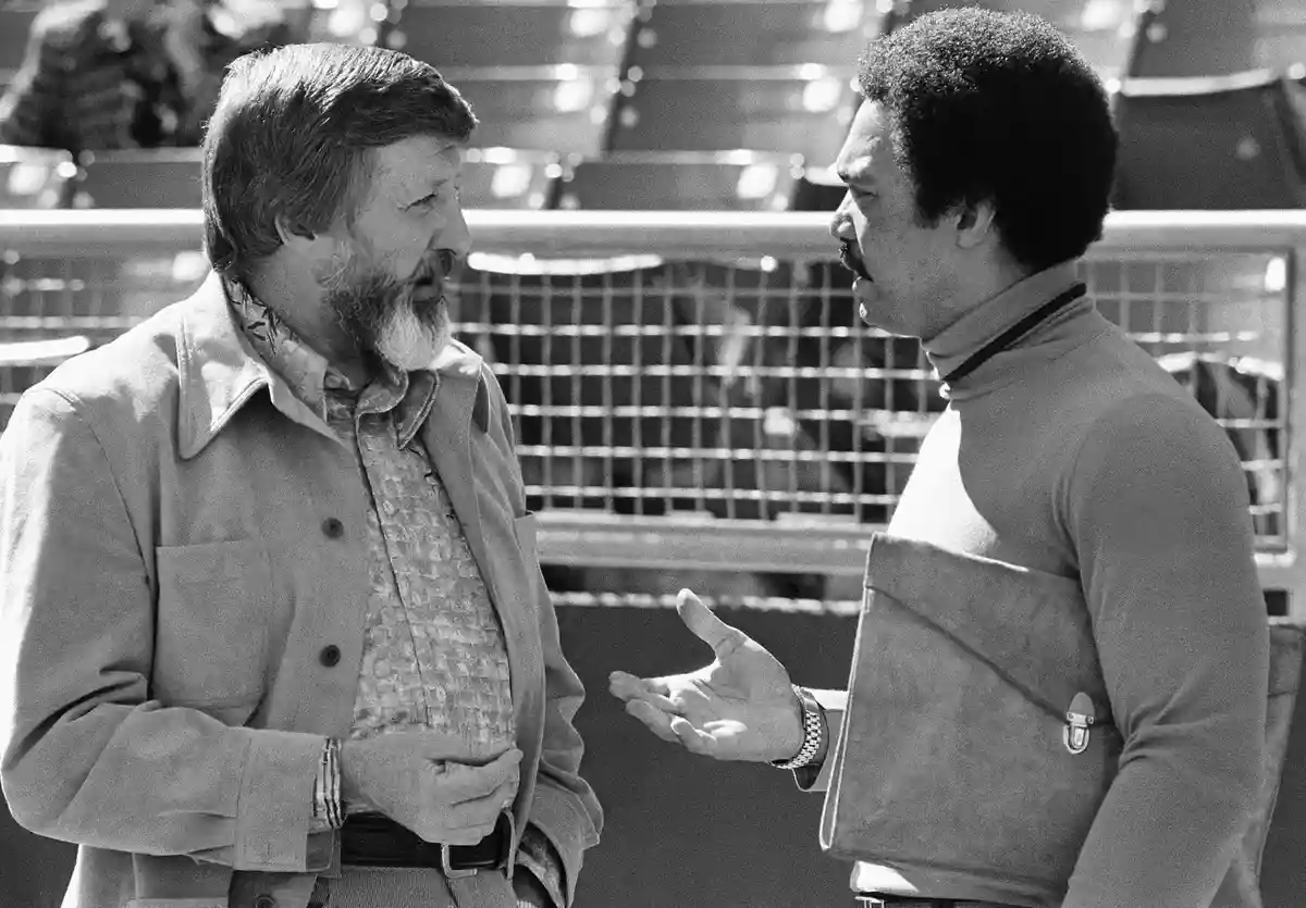 A black-and-white photo shows two men in conversation, one gesturing with his hand while the other listens. The man on the…