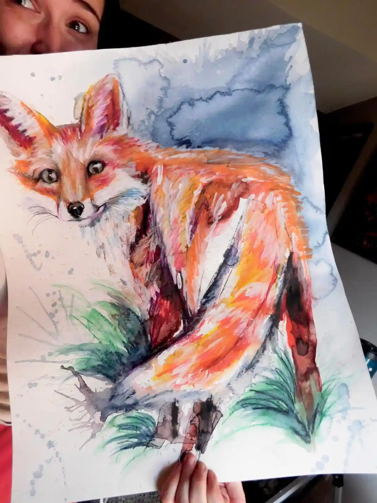 A person holds up a watercolor painting of a fox with vibrant orange, red, and white fur against a blue sky and green…