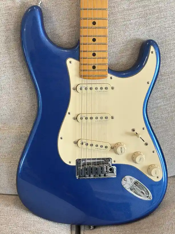 A blue Fender Stratocaster electric guitar with a maple neck and cream pickguard rests on a textured beige fabric surface…
