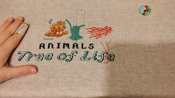 A close-up of a cross-stitch project on beige fabric showing the title 'Animals Tree of Life' with three stitched animal…