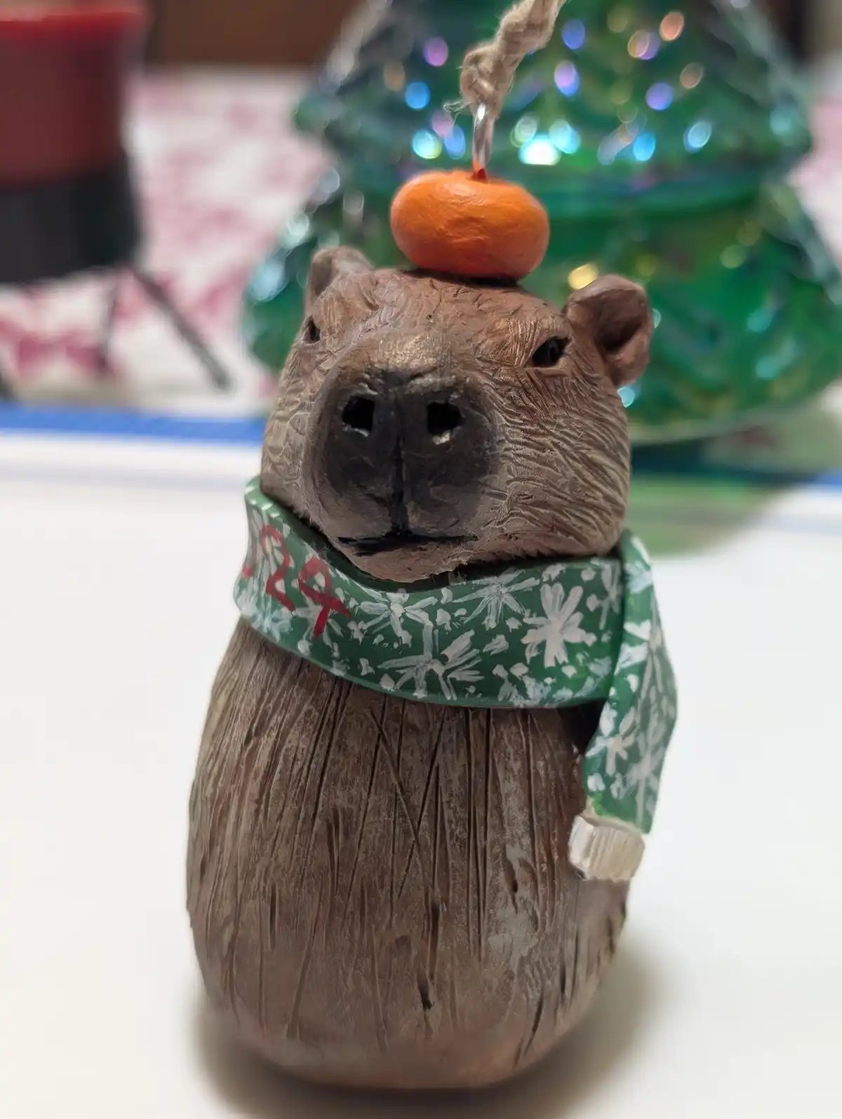 A close-up of a handmade wooden capybara ornament wearing a green snowflake-patterned scarf with '2024' written in red. A…