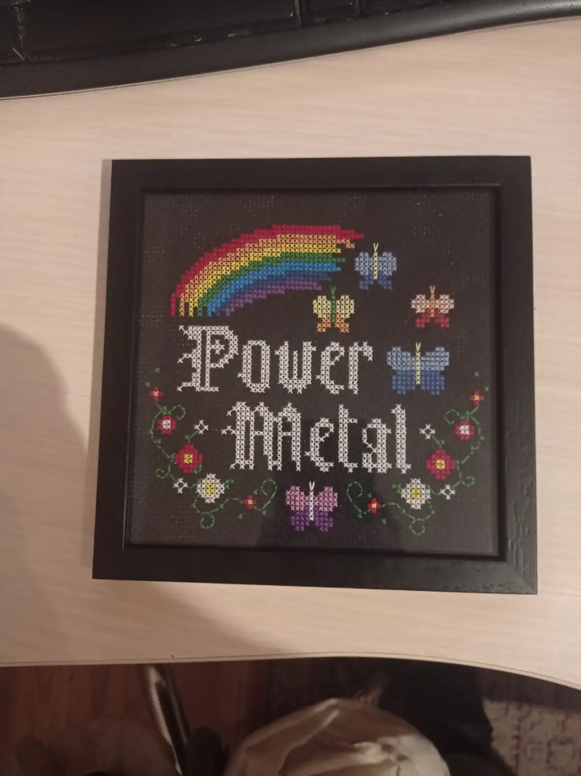 A framed cross-stitch artwork on dark fabric features a colorful rainbow, butterflies, and flowers surrounding the words…
