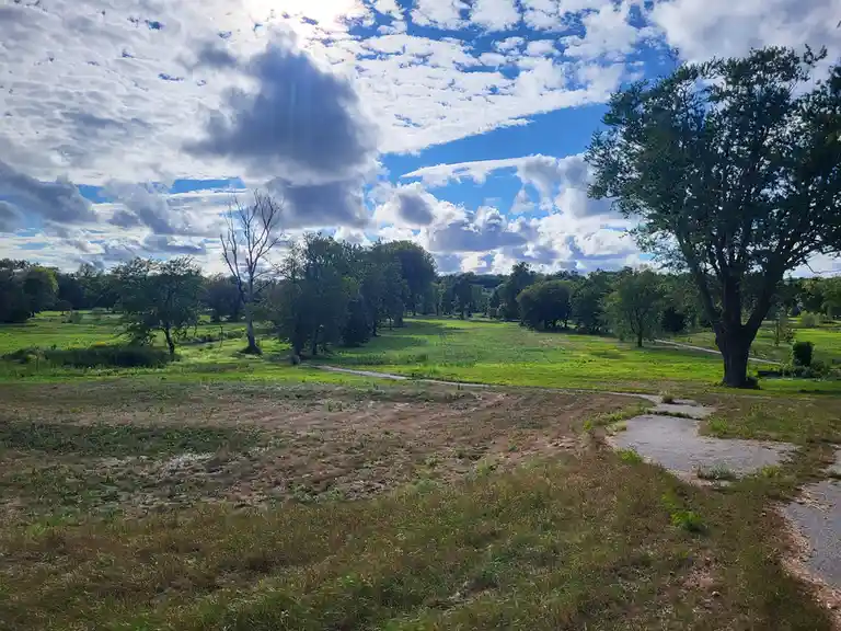 A scenic outdoor photo shows a wide grassy field with scattered trees under a partly cloudy blue sky, with a winding gravel…