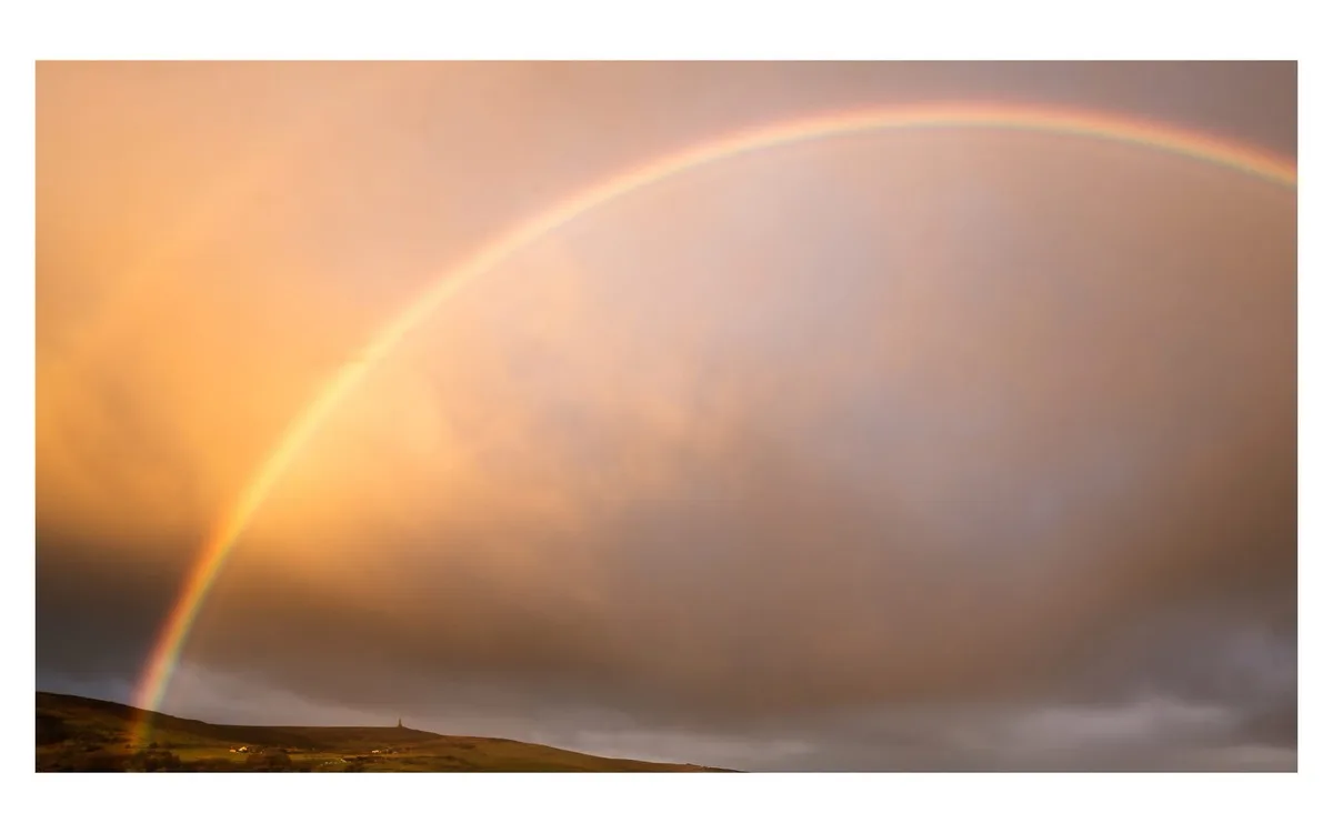 A wide-angle photograph shows a vivid, full rainbow arcing across a dramatic sky filled with orange and gray clouds over a…