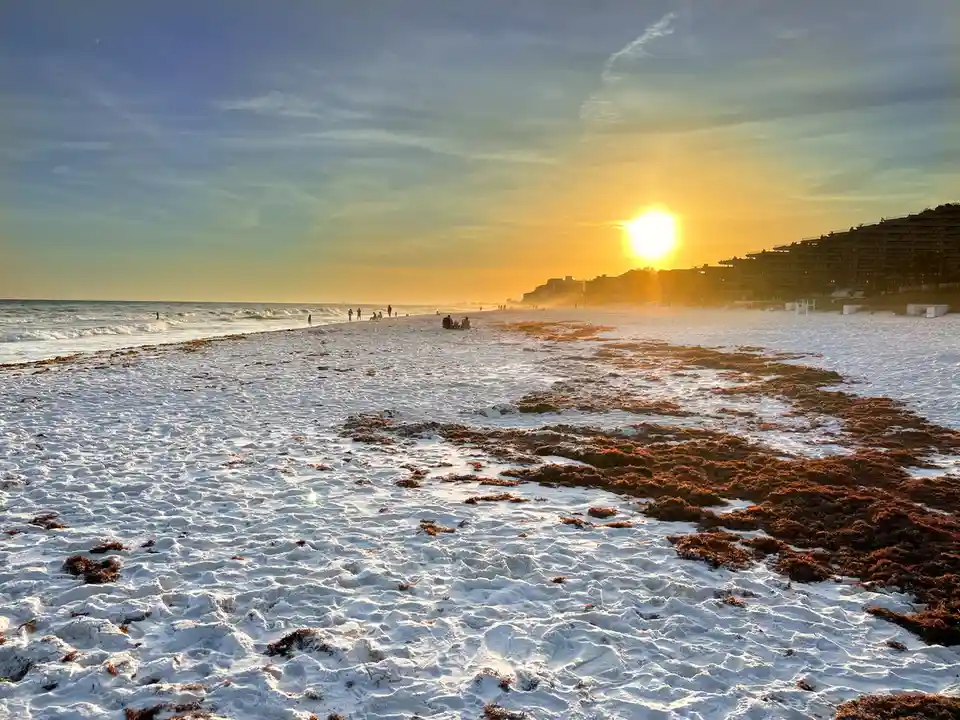 A beach scene at sunset with the sun low on the horizon, casting golden light over the ocean and distant buildings. The sand…