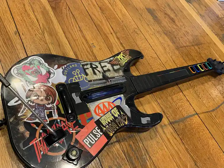 A black plastic guitar controller, likely from a music video game, lies on a wooden floor covered in numerous stickers…