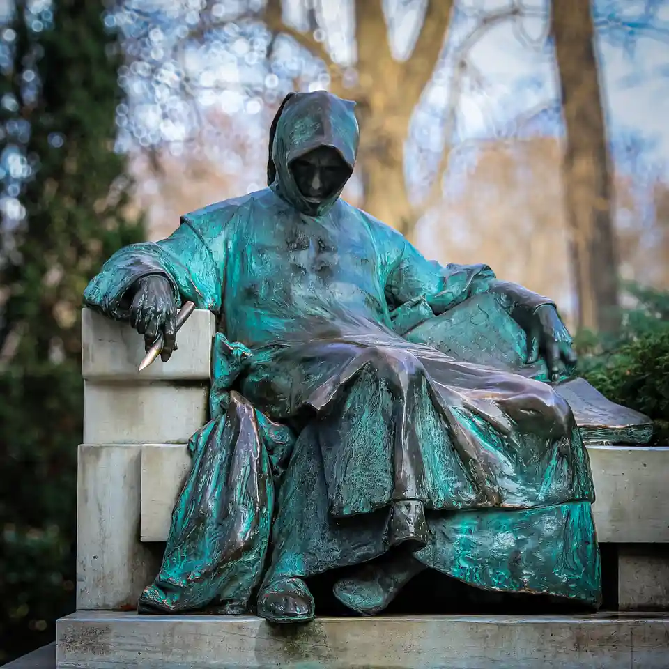 A bronze statue of a hooded figure seated on a stone bench, holding a pen and resting on an open book, with trees and sky in…