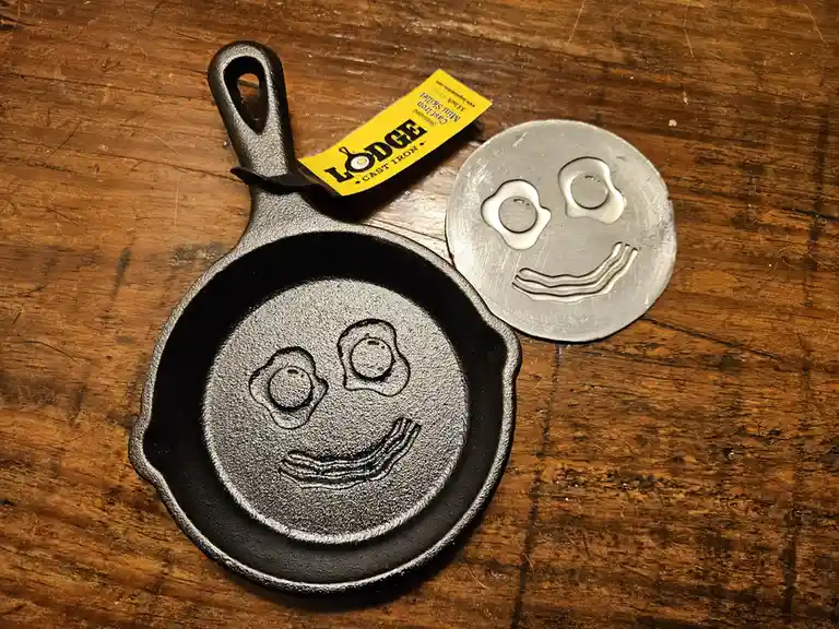 A small black Lodge cast iron skillet with a yellow tag sits on a wooden surface next to a silver metal disc. Both items…