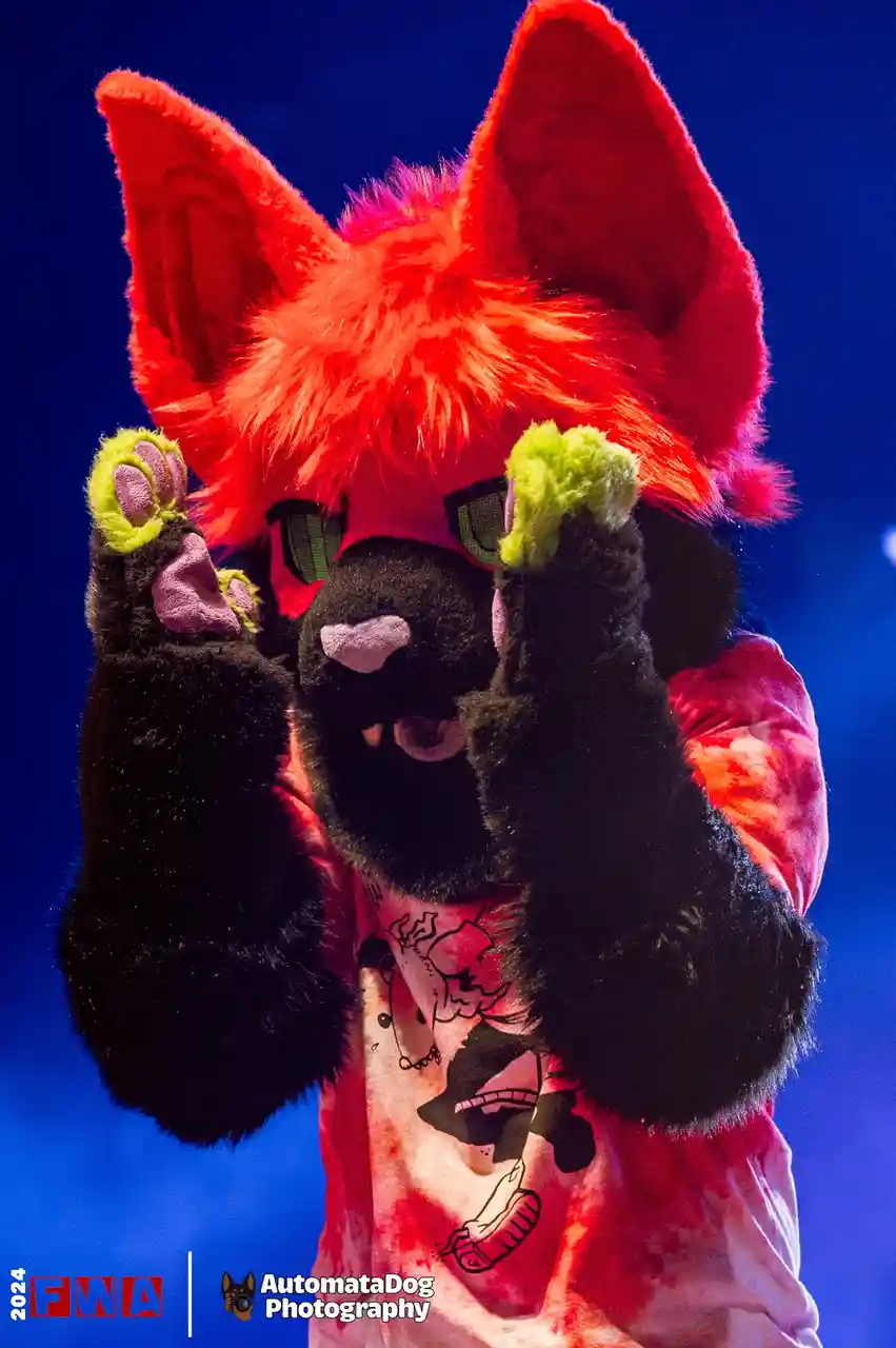 A person in a black-furred fursuit with large red ears and green-pink paw pads holds up both hands in front of their face…