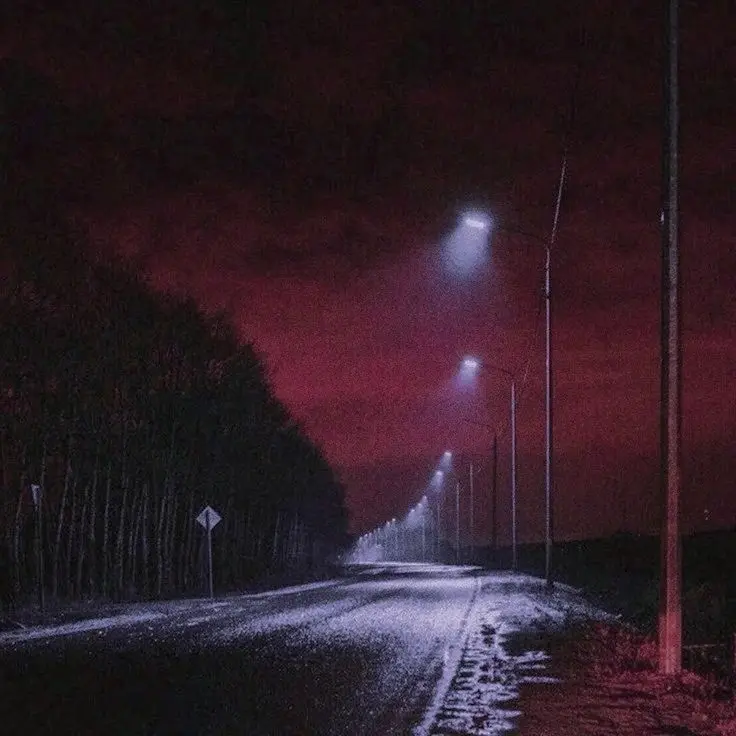 A dark, atmospheric night scene of a wet, empty road lined with streetlights receding into the distance under a deep red…