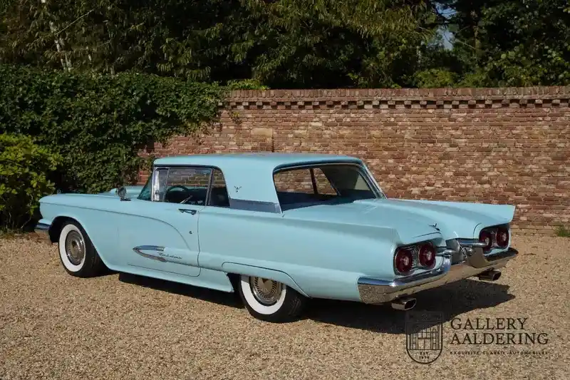 A light blue classic American car, likely a 1950s Ford Thunderbird, is parked on gravel in front of a brick wall and…