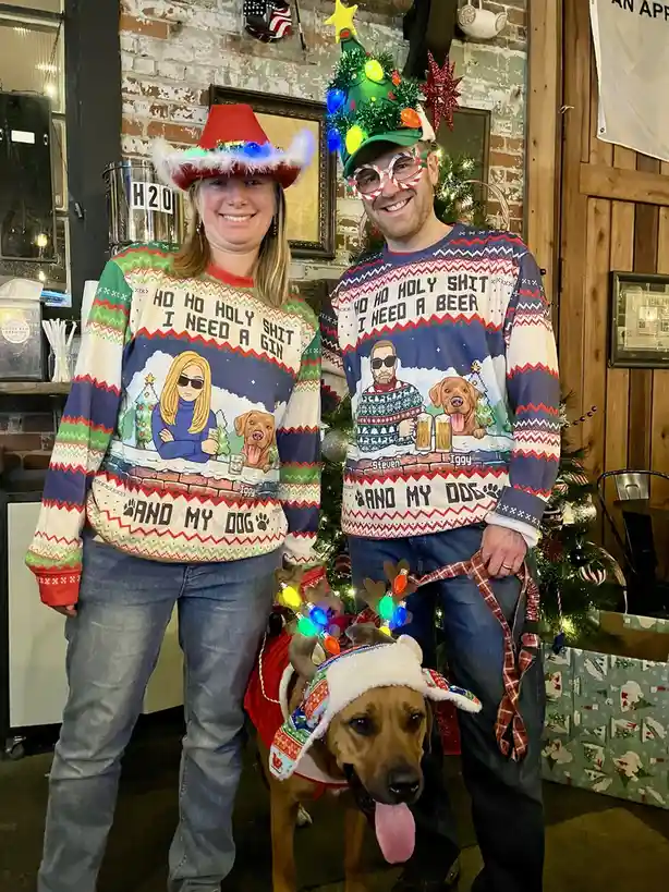 A man and woman in matching ugly Christmas sweaters stand beside their dog, all wearing festive holiday hats and…