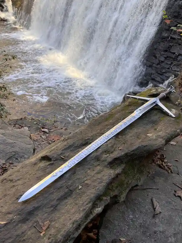 A silver sword with a crossguard and engraved blade rests on a mossy rock beside a cascading waterfall, with the overlaid…