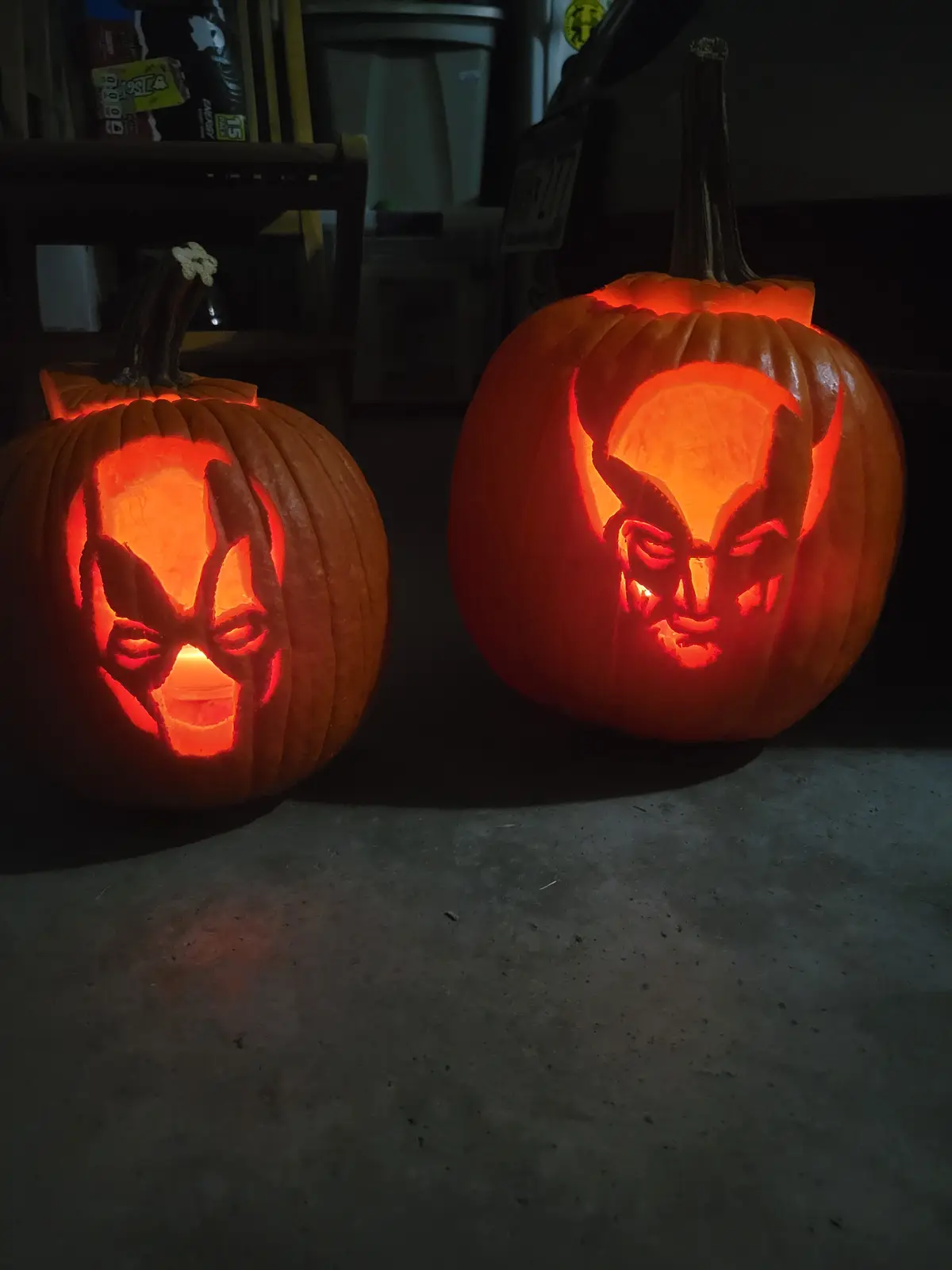 Two intricately carved Halloween pumpkins sit side by side on a concrete floor, illuminated from within by a warm orange…