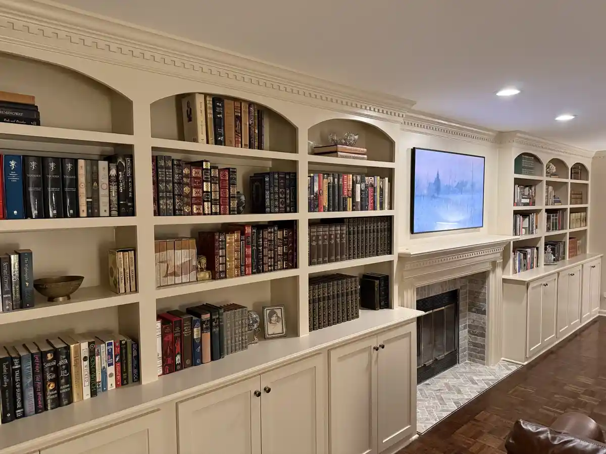 An image of a luxurious, well-lit home library with white built-in bookshelves filled with numerous books, a fireplace, and…