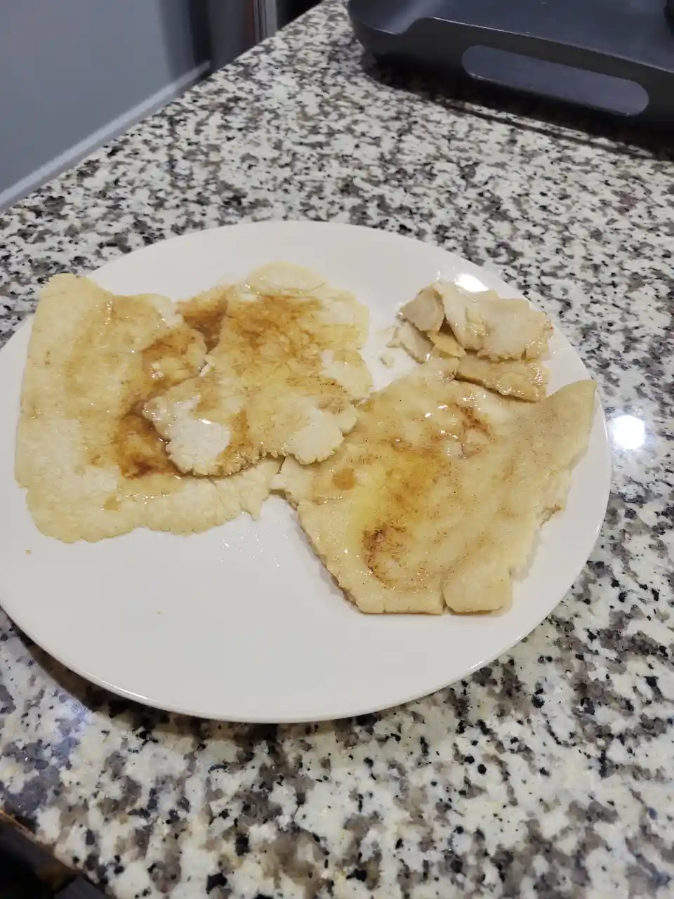 A white plate holds several lumpy, unevenly cooked, pale yellow pancake-like items covered in syrup and cinnamon, sitting on…
