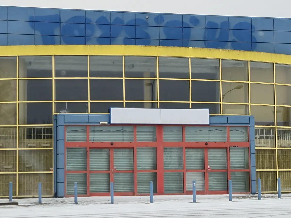 The image shows the exterior of a closed Toys R Us store with its iconic blue and yellow facade, red doors covered by…