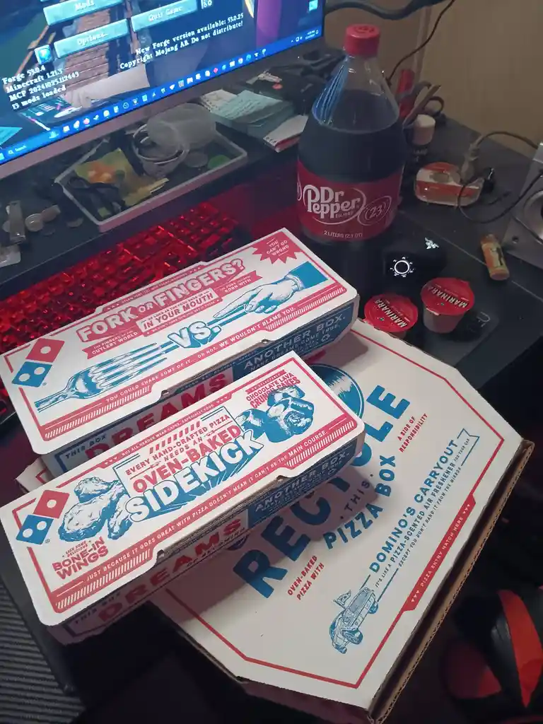 A photo of a cluttered desk with a computer monitor showing Minecraft, a red-lit keyboard, and a 2-liter Dr Pepper bottle…