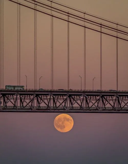 A large, orange full moon is positioned directly beneath a suspension bridge at dusk, appearing to be resting on the water…