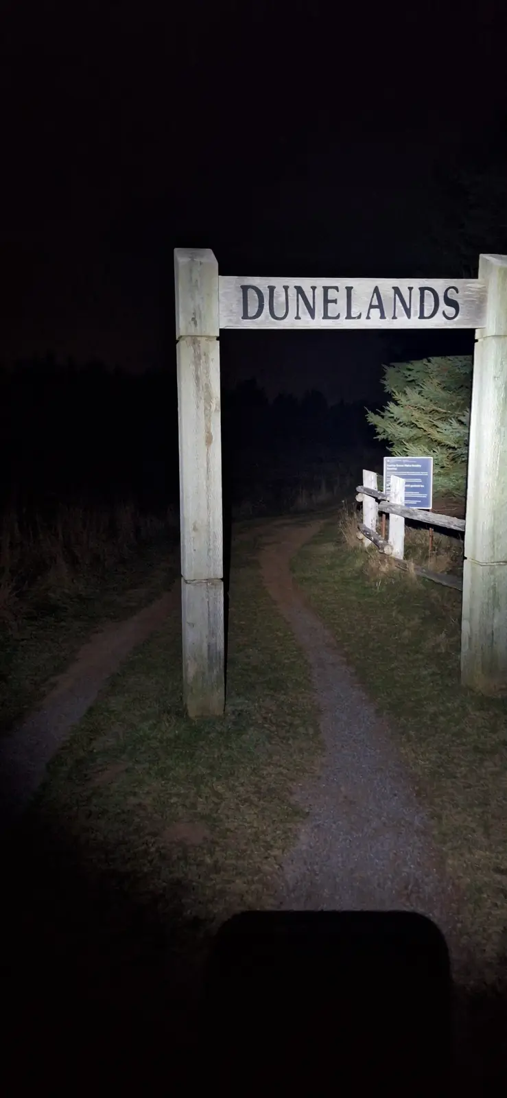 A nighttime photo shows a wooden archway sign reading 'DUNELANDS' over a dirt path leading into darkness, with a fence and…