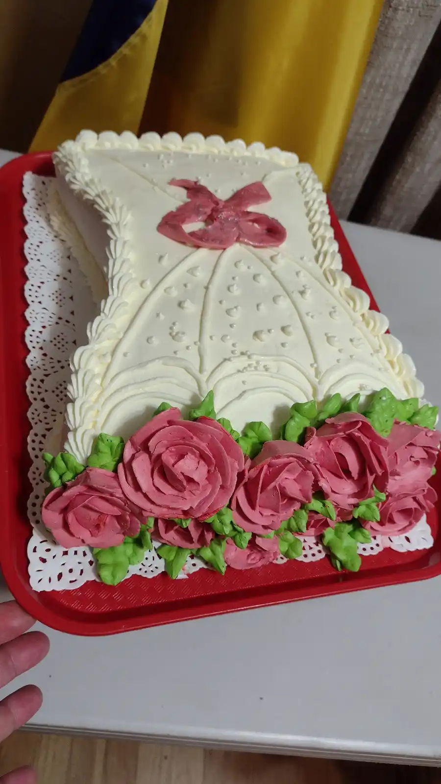 A rectangular cake decorated to resemble a white dress with piped frosting details, a pink bow at the top, and pink roses…