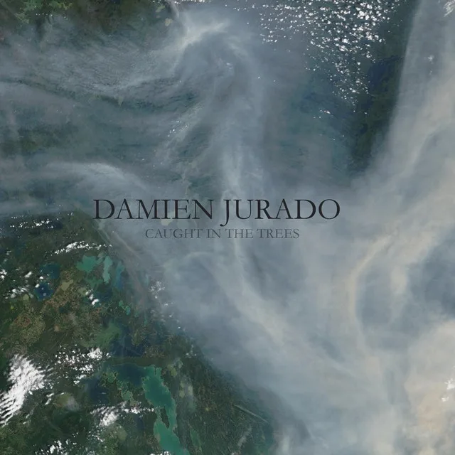 An aerial satellite image shows swirling clouds and green landmasses, overlaid with the text 'DAMIEN JURADO' and 'CAUGHT IN…