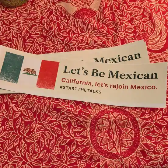 Two stickers lie on a red patterned fabric, featuring a hybrid flag with California's bear and Mexico's green, white, and…