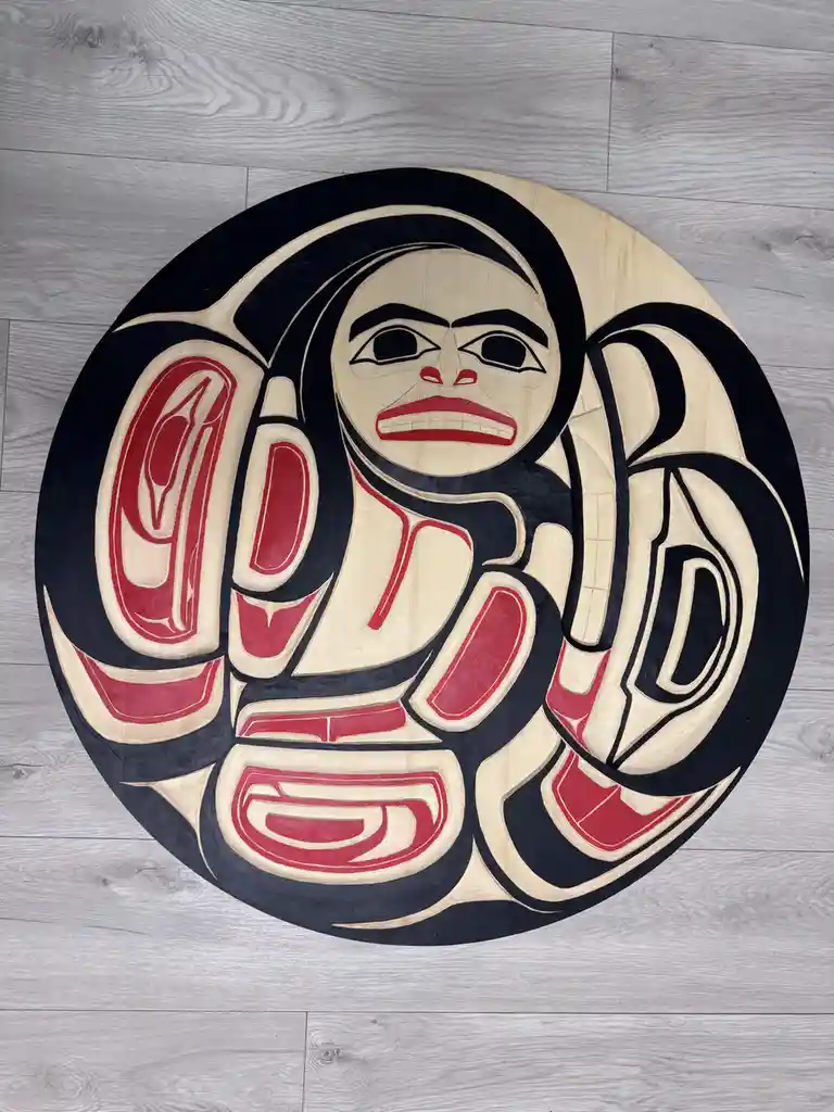 A circular wooden carving with a stylized face and abstract patterns in black, red, and natural wood tones, resembling…