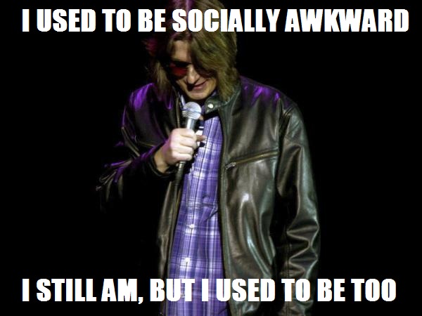 A man with long hair and red sunglasses, wearing a leather jacket over a plaid shirt, holds a microphone on a dark stage…