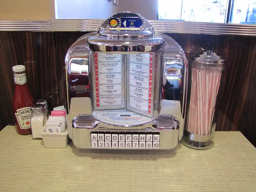 A vintage chrome jukebox sits on a diner counter next to a Heinz ketchup bottle, salt and pepper shakers, sugar packets, and…