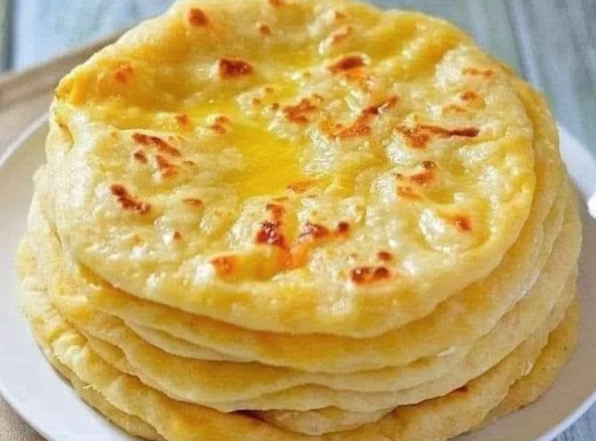 A stack of golden-brown, buttery flatbreads sits on a white plate, glistening with melted butter and showing slight charred…